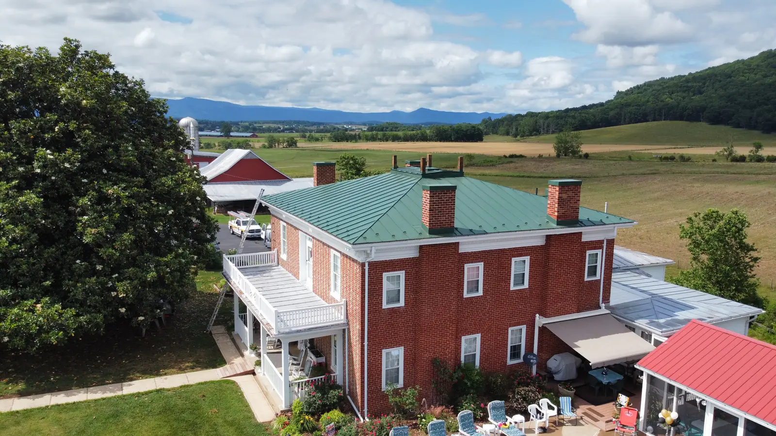 A red brick farmhouse under a green standing-seam metal roof, with fields and the Blue Ridge behind