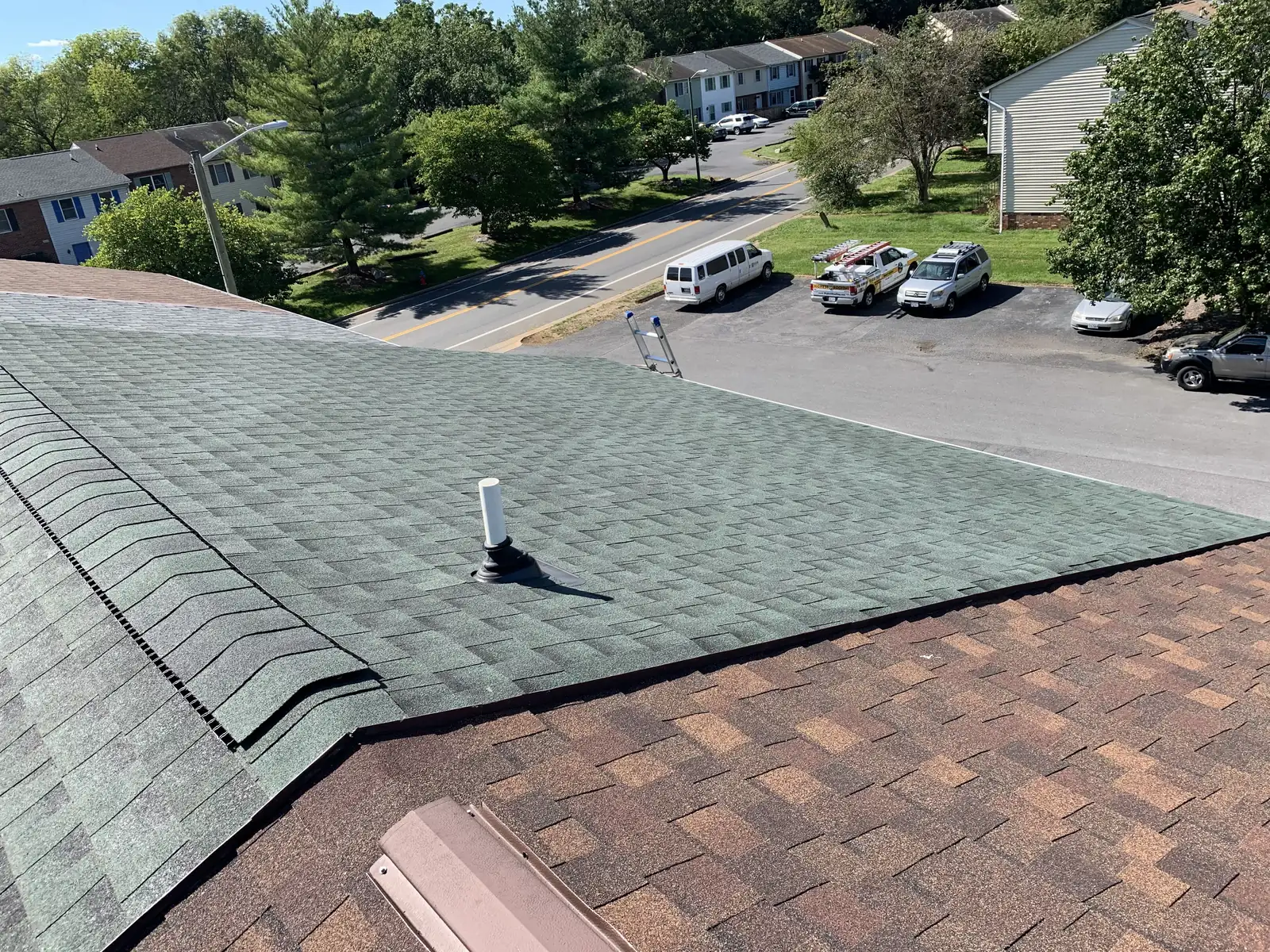A green shingle roof being replaced next to an older brown roof, with the crew truck and ladders below