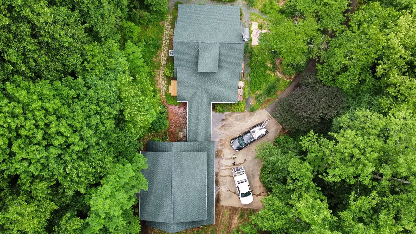 A house with a green-gray shingle roof among trees, seen from straight above, with trucks in the drive