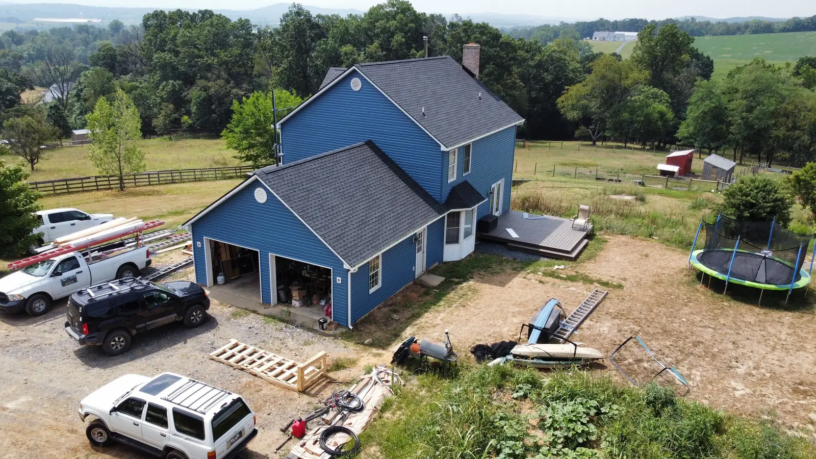 Highest Roofing trucks and ladders at a blue farmhouse job site, with fields and hills behind