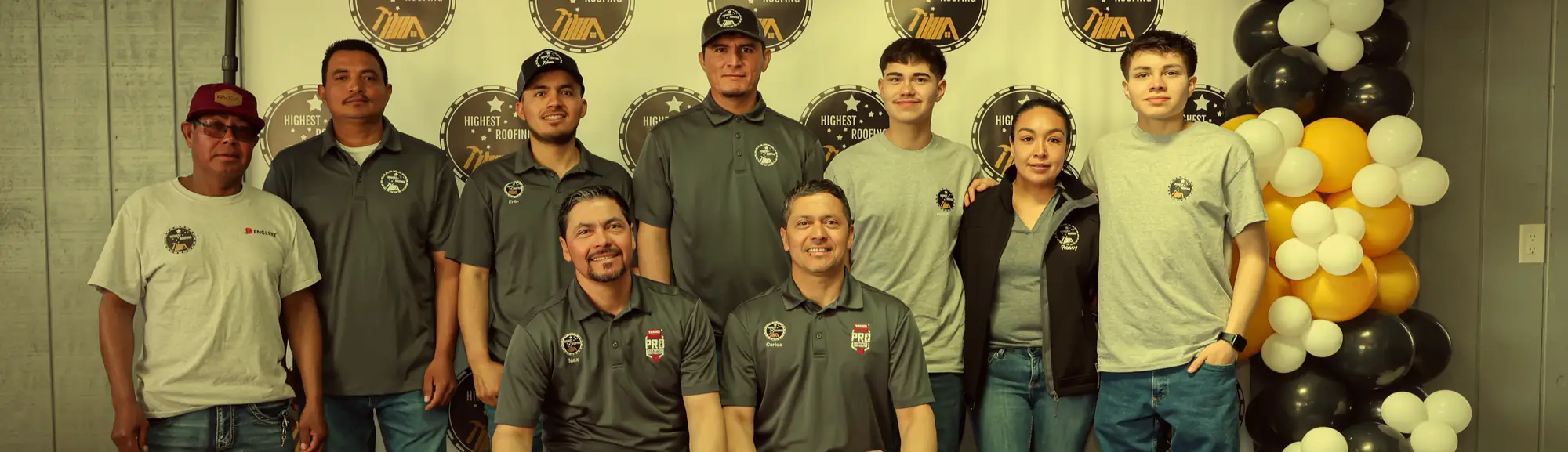 The Highest Roofing team in company shirts in front of a logo backdrop, with Carlos and Max seated in front